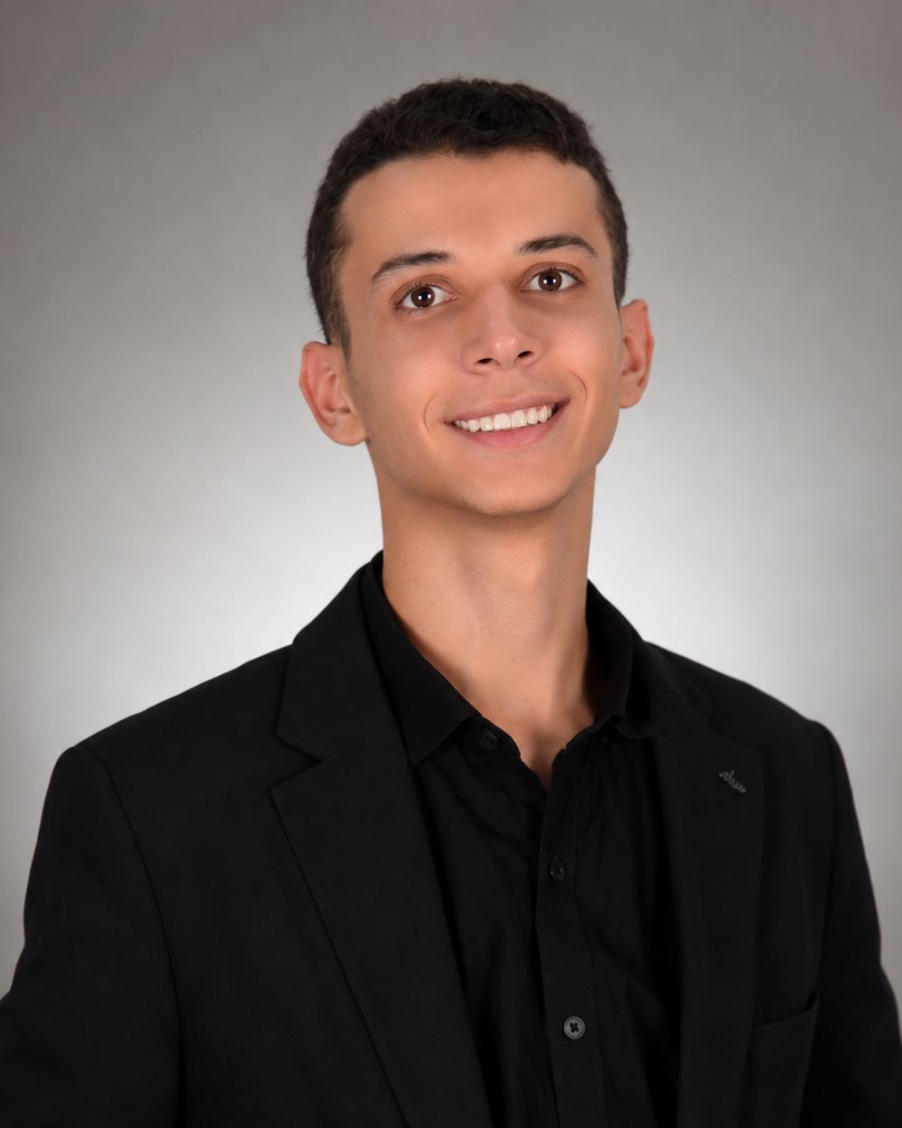 Yousef smiling in a black suit against a neutral grey background.